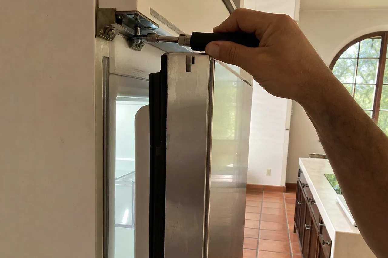 Hand turning a black-handled nut driver on the upper hinge bracket of an open stainless built-in refrigerator door, with terracotta floor tiles and an arched window behind